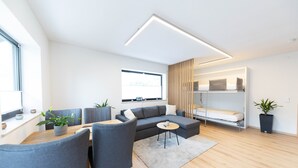 Una sala de estar moderna con un sofá, mesa de comedor, sillas, una pequeña mesa de centro, una planta en maceta, una ventana y una cama empotrada con una mesita de noche.