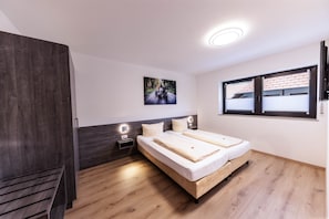 A bedroom with two beds, a dark wardrobe, a nightstand with a lamp, a picture on the wall, and a window with blinds.