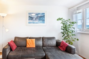 A dark leather sofa with red and orange cushions, a framed painting of a winter landscape, a floor lamp, and a potted plant.