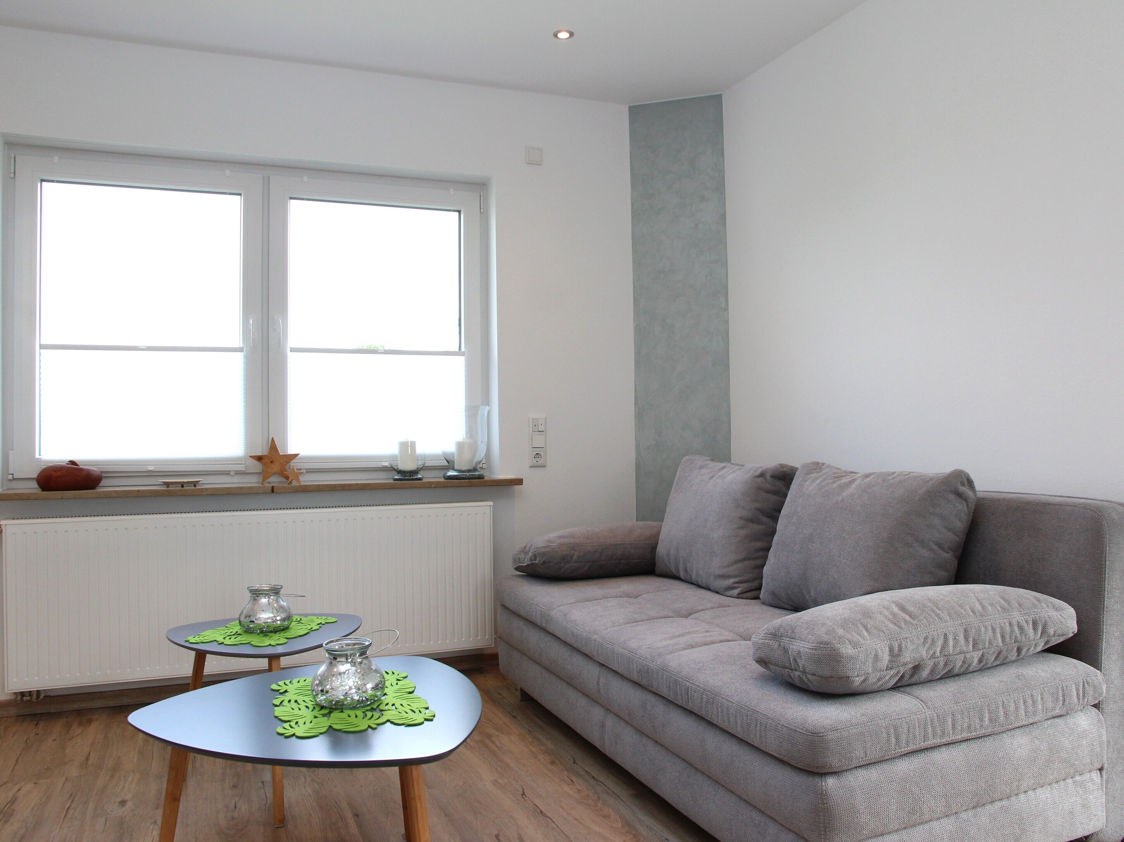 A living room with a grey sofa, a small round table with a glass vase, a window with blinds, and a radiator.