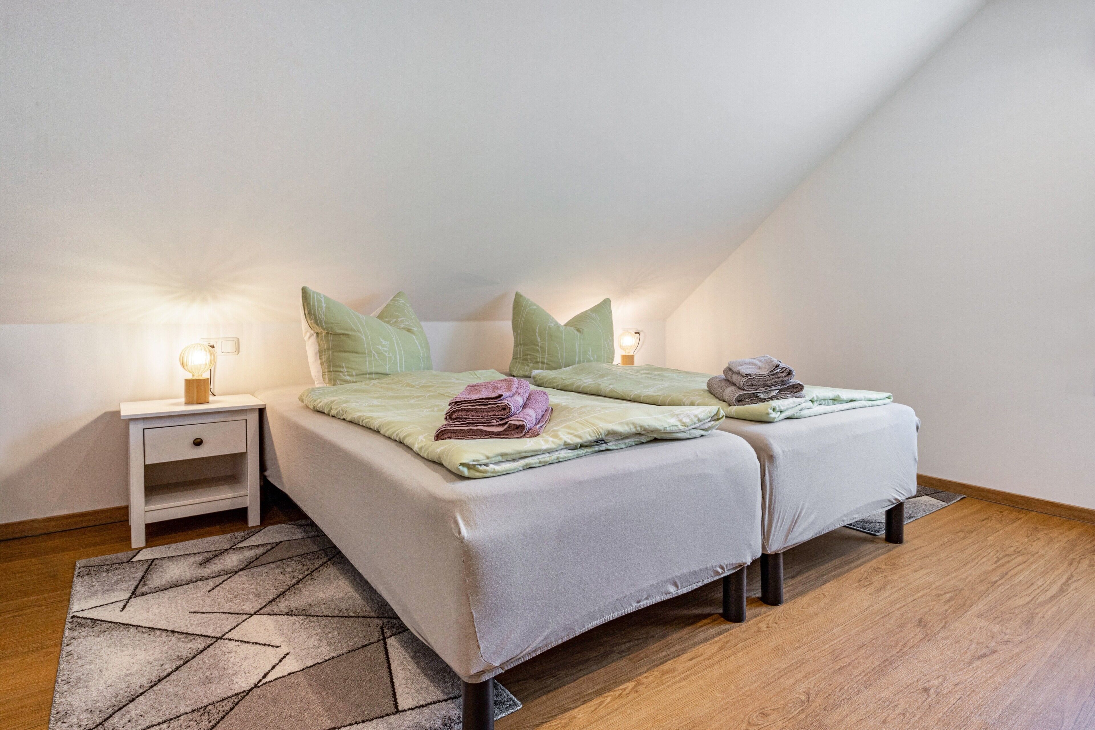 Ein Schlafzimmer mit Bett, Kissen, Nachttisch, Lampe und einem gemusterten Teppich.