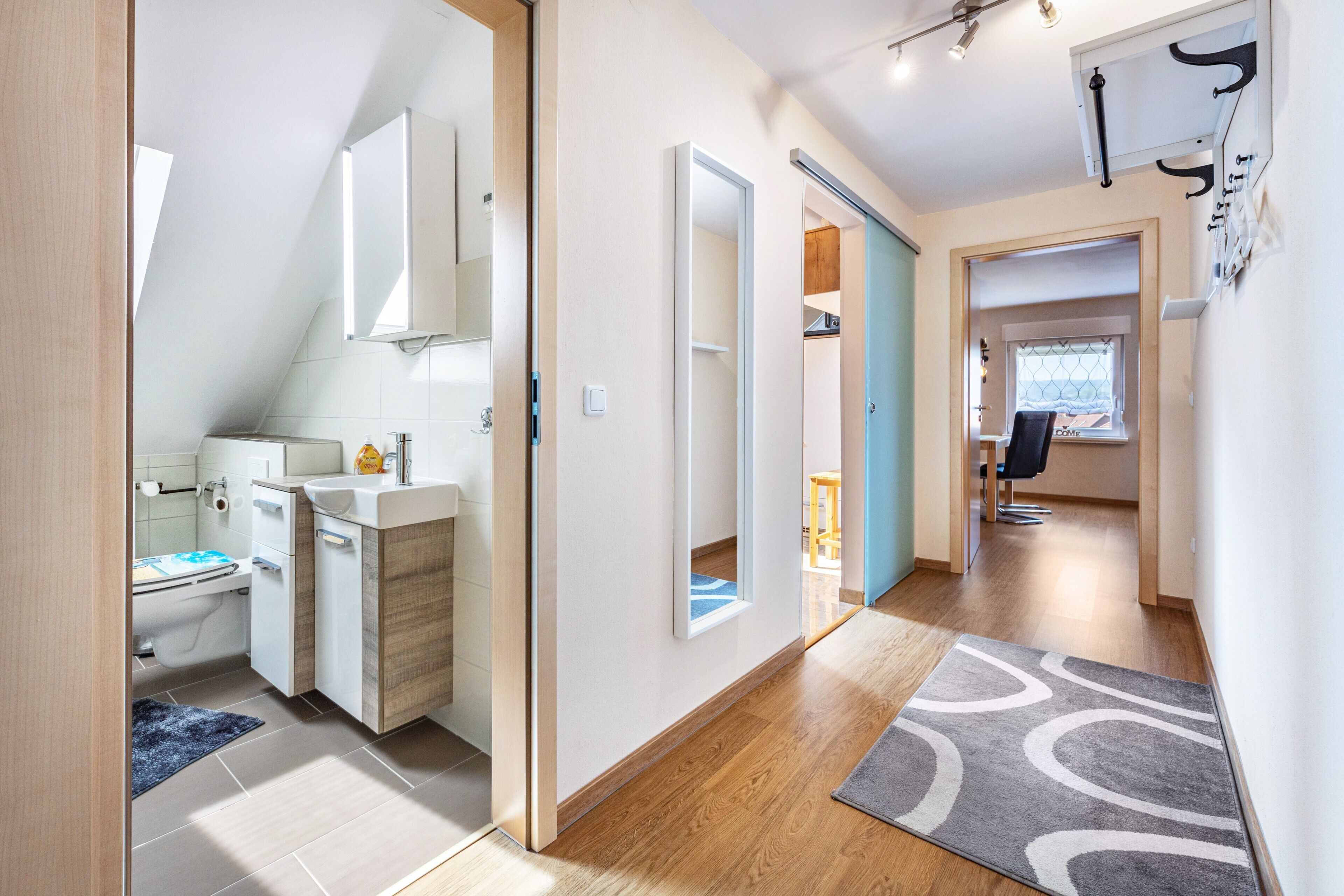 A modern interior with a bathroom, a hallway leading to a dining area, and a visible window.