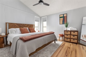 Un dormitorio con cama de madera, ventilador de techo, mesita de noche, lámpara de noche, televisor montado en la pared, ventana y aparador de madera con cestas.