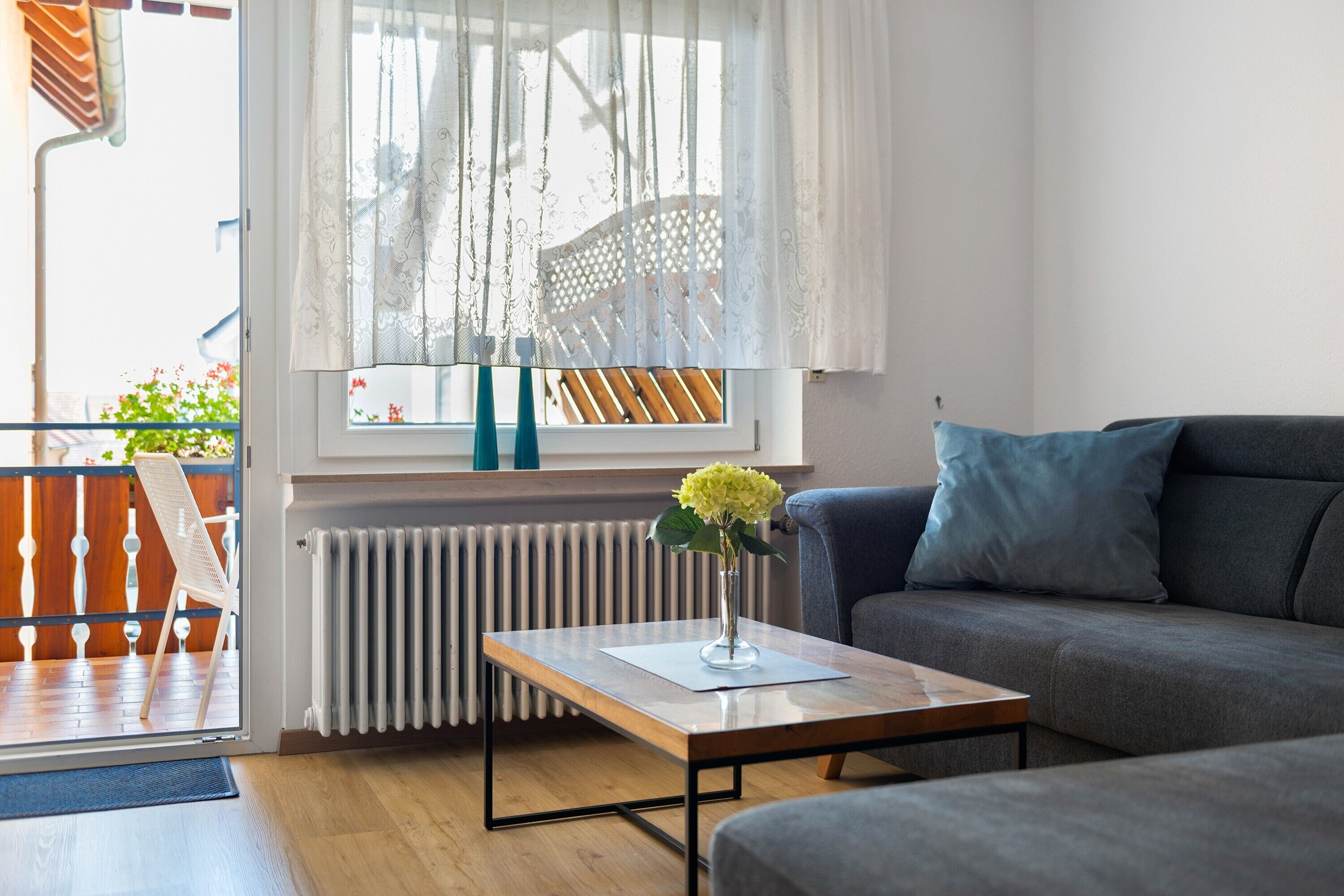 Un salon moderne avec un canapé, une table basse ornée d’un vase de fleurs, un radiateur et une vue sur une terrasse en bois.