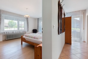 Ein Schlafzimmer mit Bett, Heizkörper, einem Fenster mit durchsichtigen Vorhängen, einer Pendelleuchte und einer Holztür.