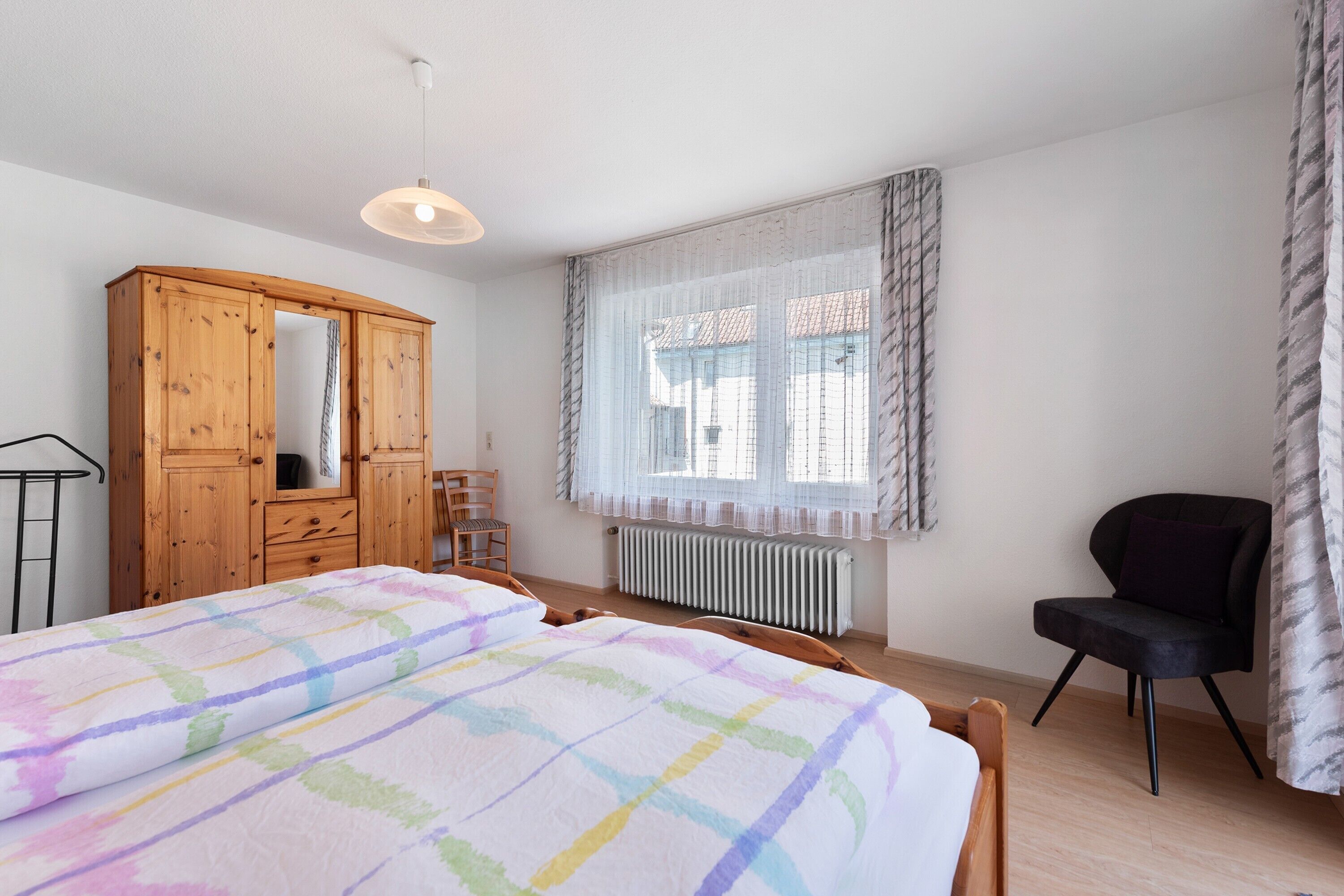 A bedroom with a bed, wooden wardrobe, chair, and a window with curtains.