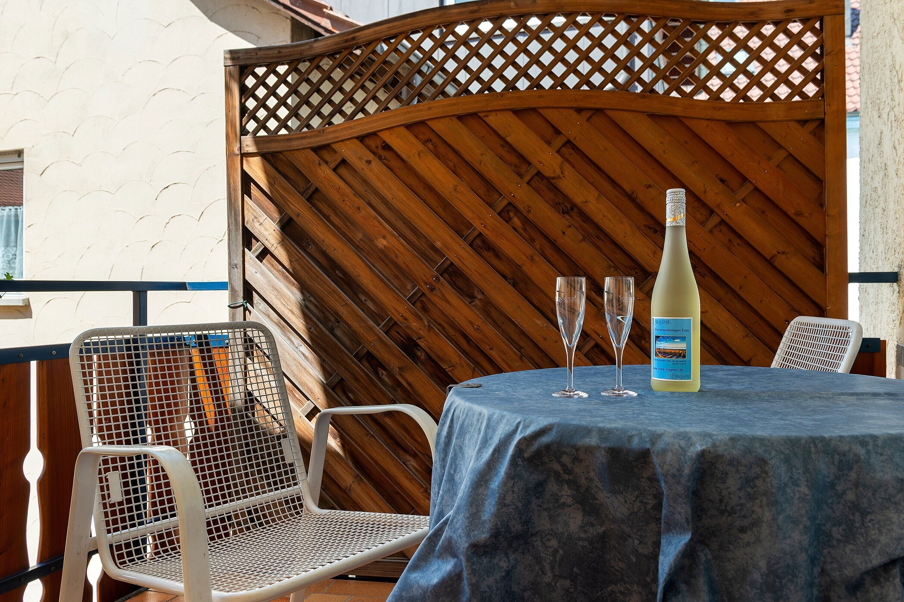 A table with a blue tablecloth, two wine glasses, and a bottle of wine on a balcony with metal chairs and a wooden lattice partition.