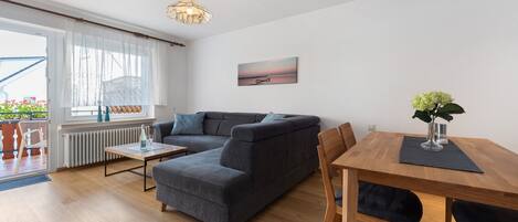 Una sala de estar moderna con un sofá seccional gris, una mesa de comedor de madera con sillas, una mesita auxiliar, una lámpara colgante y un cuadro en la pared.