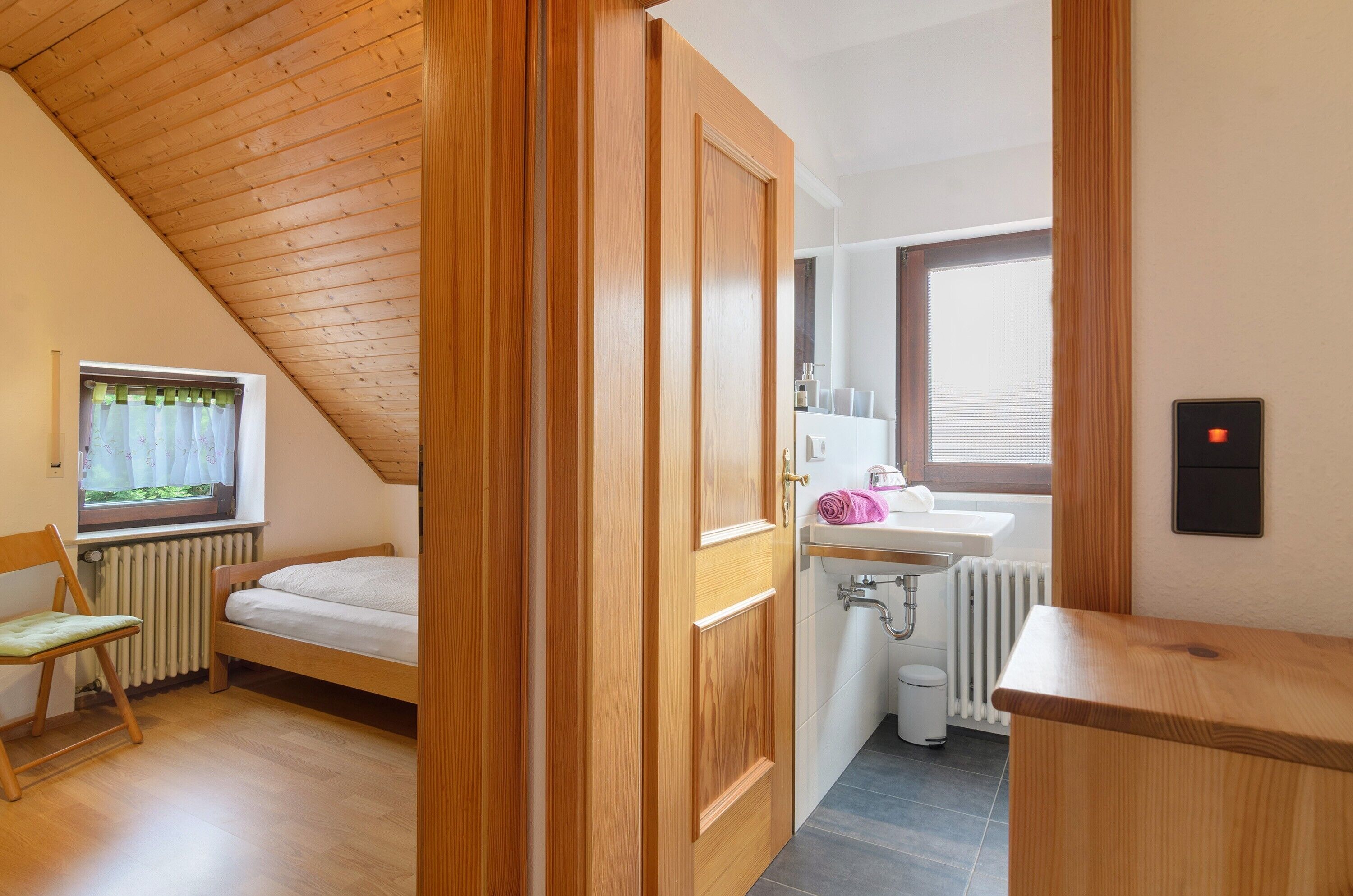 Camera da letto con soffitto in legno, un letto, una sedia, una finestra, un termosifone, un lavandino e uno specchio.