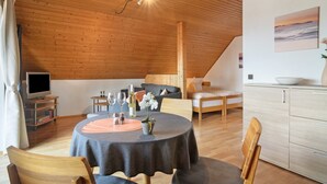 Ein gemütliches Zimmer mit Holzbalkendecke, einem Esstisch mit Stühlen, einer Couch, einem Fernseher auf einem Schrank und einer Küchenzeile mit Schränken.