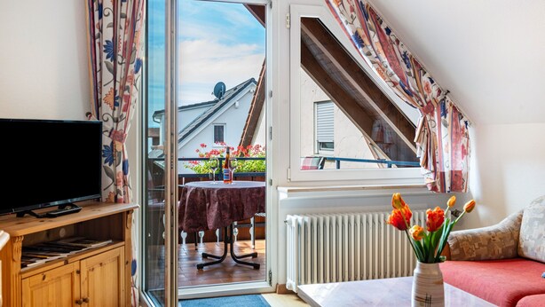 A living room with a television, a table with a vase of flowers, a sofa, and a view of the outside. - Apartment '3' with Private Garden, Balcony and Wi-Fi (Hagnau am Bodensee)