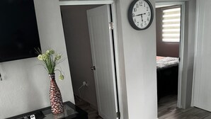 Una habitación con piso de madera, una mesa negra con un florero, un reloj de pared y una puerta que da a otra habitación.
