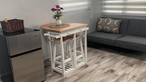 Un espacio habitable compacto con kitchenette, una pequeña mesa de comedor, un sofá y un ventanal con persianas.