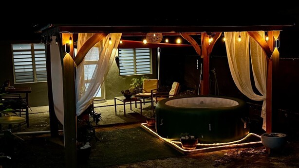 Un jacuzzi bajo una pérgola con luces colgantes, rodeado de sillas y una mesa.