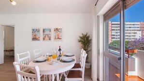 Un comedor con una mesa redonda, sillas blancas y vistas a edificios a través de un amplio ventanal.