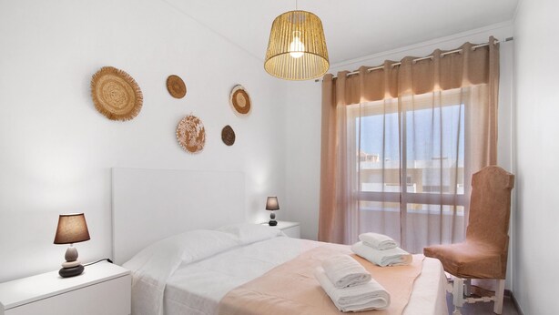 Una habitación de hotel con una cama, una silla, una mesita de noche, una lámpara y un ventanal con cortinas.