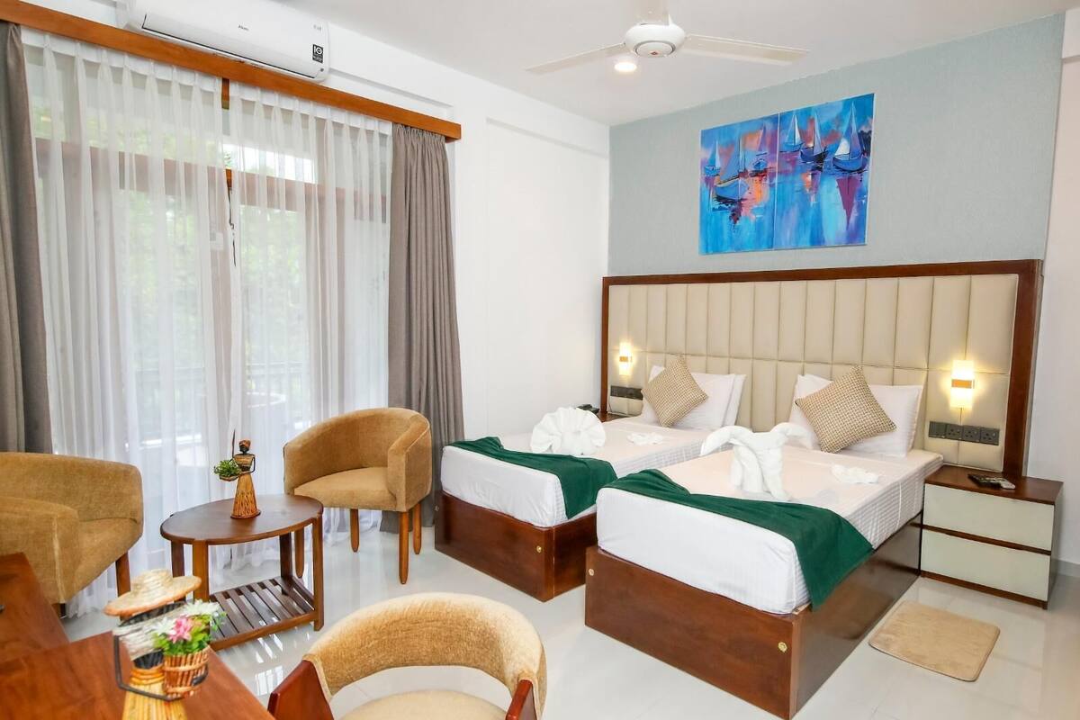 A hotel room with two beds, a sitting area with a chair and table, a ceiling fan, and a painting on the wall.