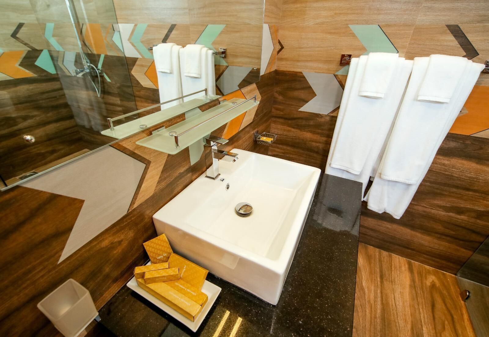 A modern bathroom with a large white sink, a glass shower enclosure, and a mirror. There are multiple white towels and a soap dish on the counter.