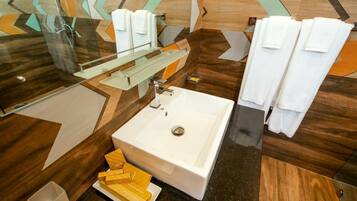 A modern bathroom with a large white sink, a glass shower enclosure, and a mirror. There are multiple white towels and a soap dish on the counter.