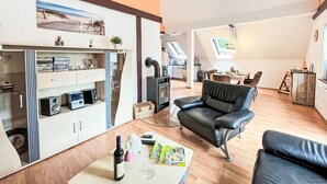 A modern living room with a black leather sofa, a dining area with a table and chairs, a kitchen in the background, and a large window providing natural light. - Cosy apartment in villa for 8 people with WIFI, TV, terrace and pets allowed (Bad Wildungen)