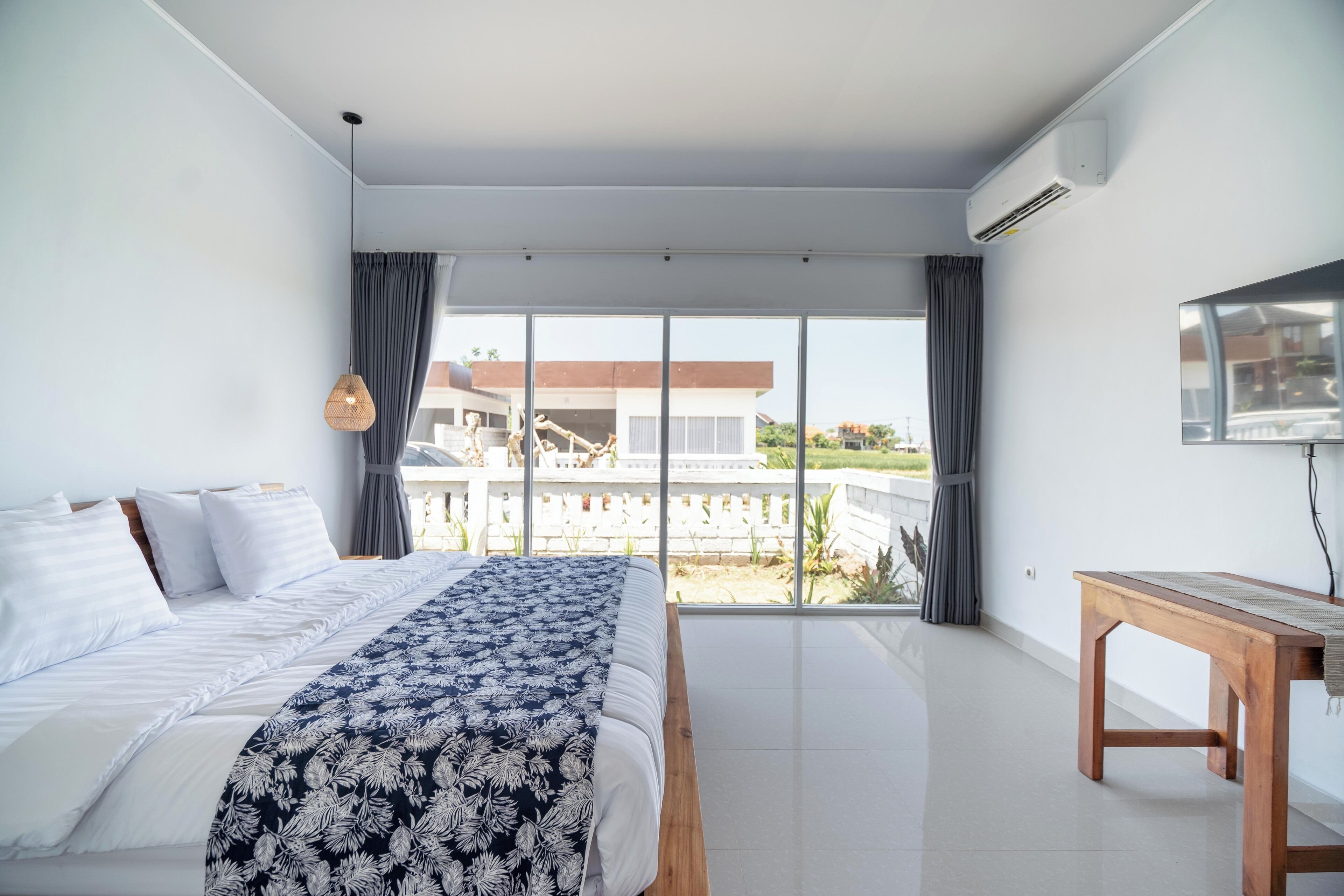 Un dormitorio moderno con una cama grande, una mesita de noche de madera y vistas a un edificio a través de amplios ventanales.
