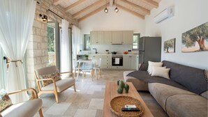 Een ruime woonkamer met een bank, stoelen en een houten salontafel. Achtergrond toont een keuken met kasten en een koelkast.