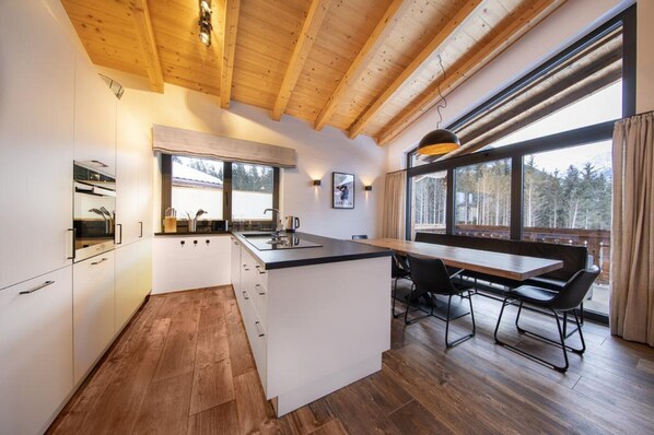 A modern kitchen with wooden beams, a large window, and a dining area with a table and chairs. - Chalet in Neukirchen by the Slopes (Neukirchen am Grossvenediger)