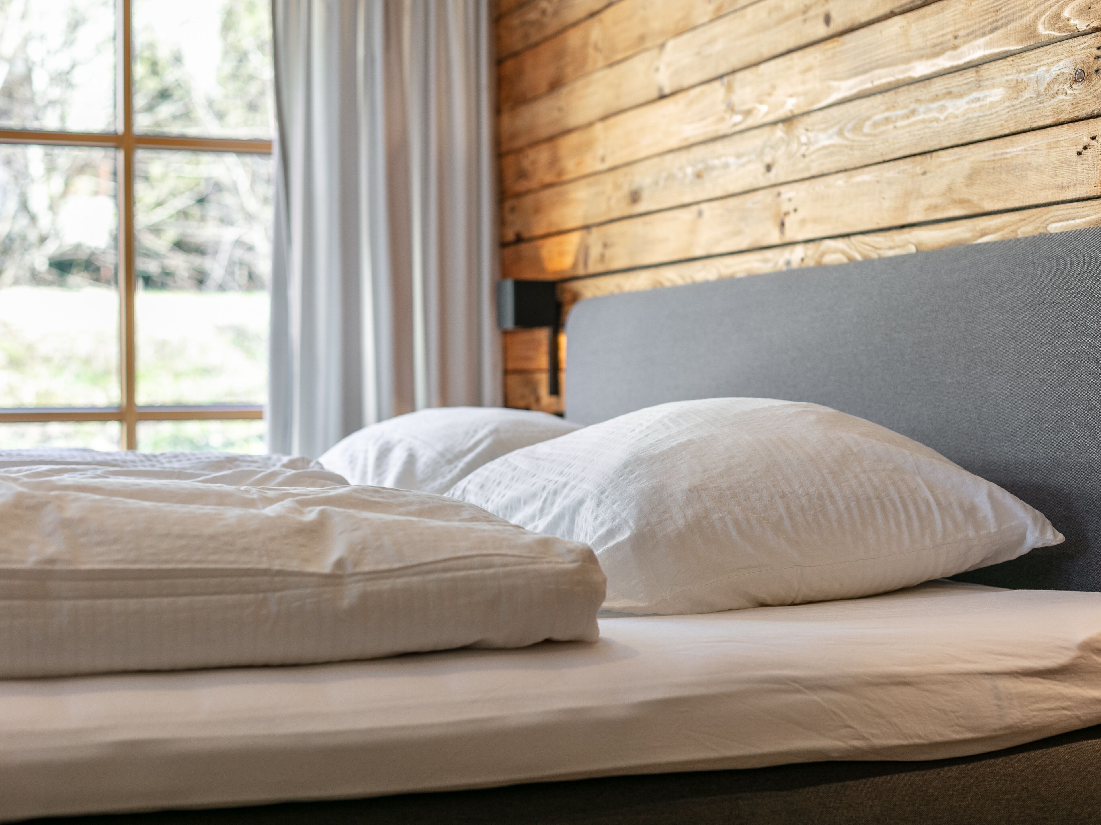 A bed with white bedding and pillows, a grey headboard, and a window with white curtains.