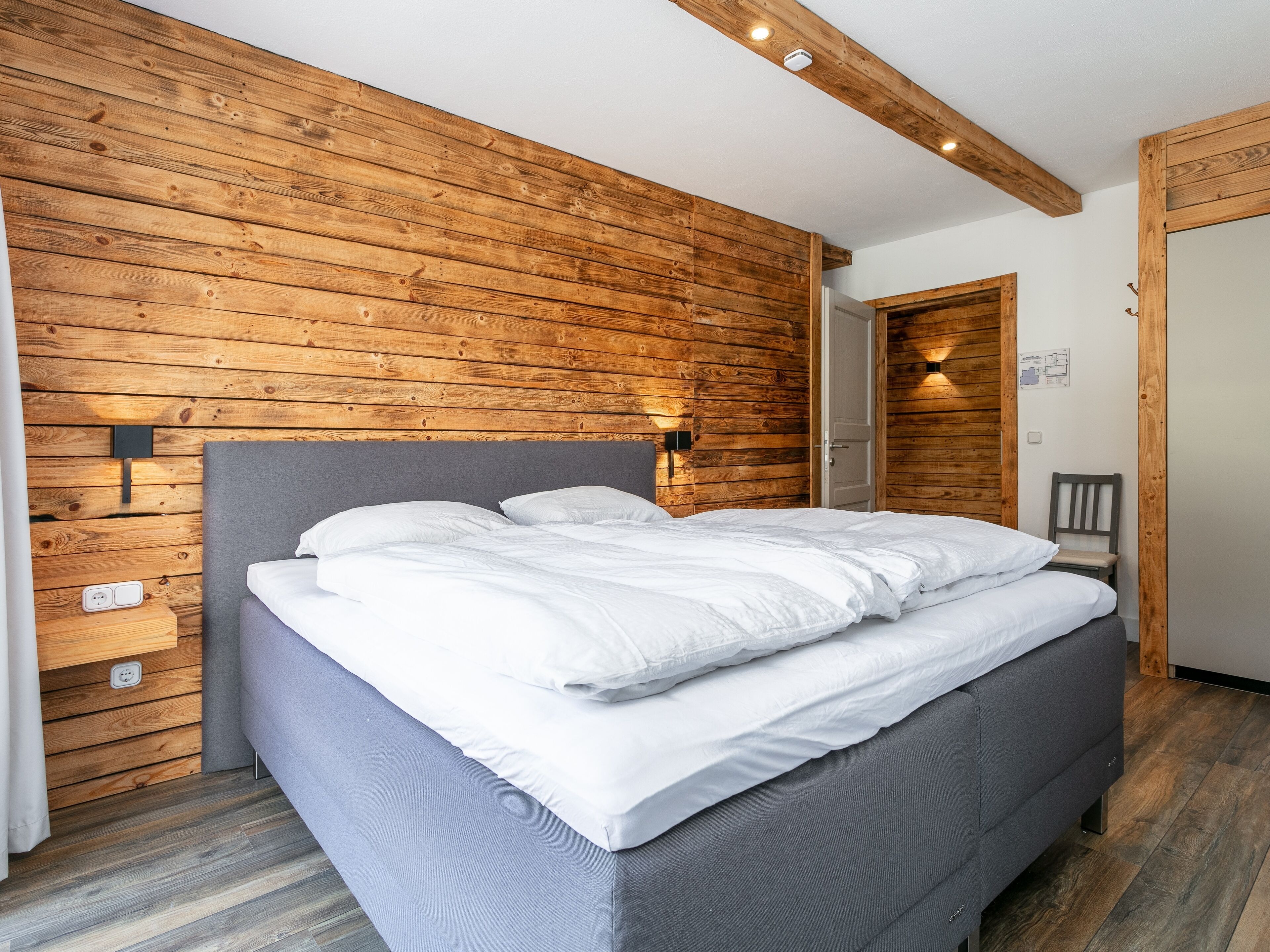 Une chambre avec un mur en bois, un lit avec une literie blanche, une tête de lit grise et une table de chevet dotée d’une prise électrique.