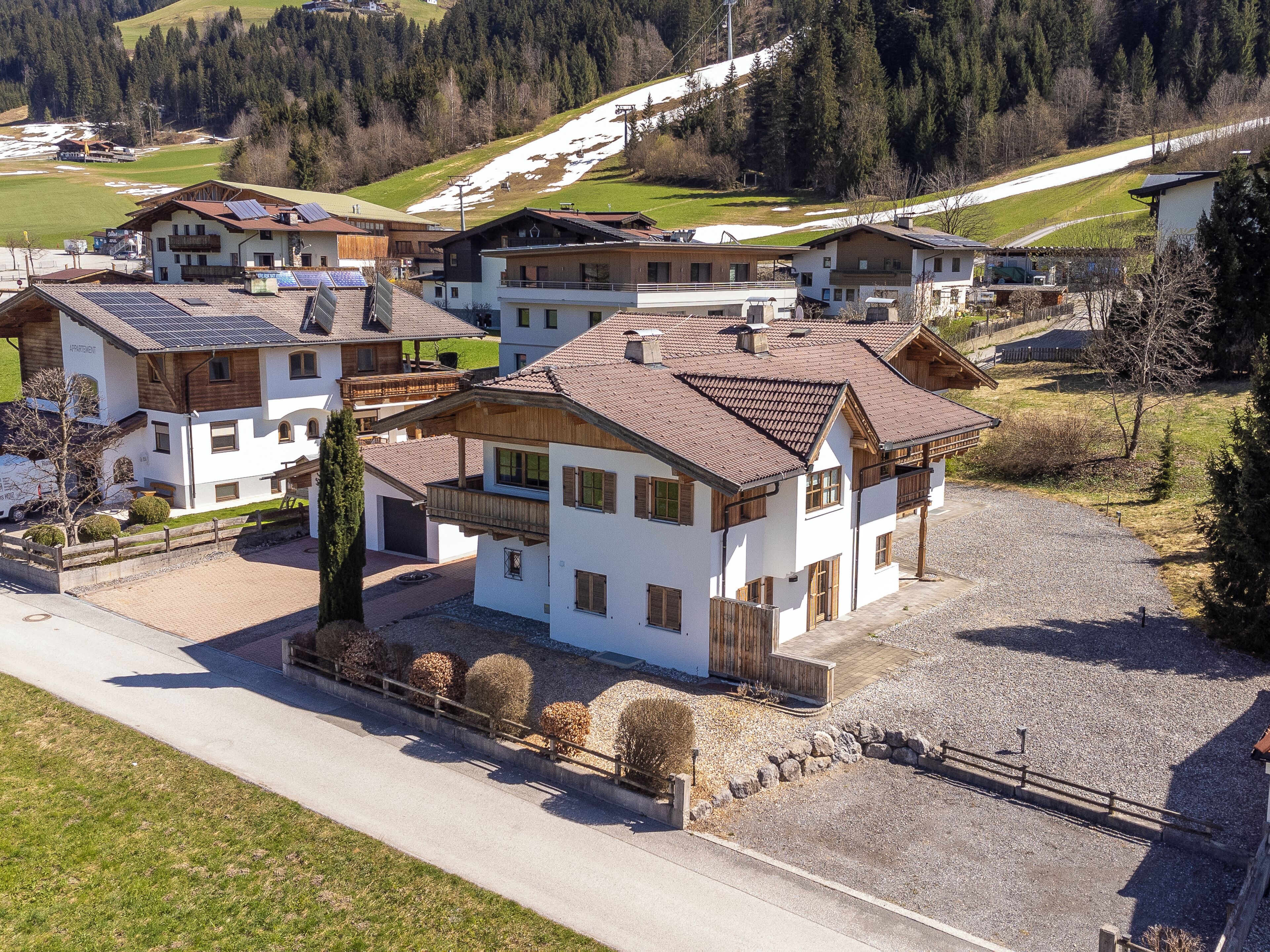Zona residenziale con diverse case, alcune dotate di pannelli solari, circondata da campi verdi e un pendio boscoso.