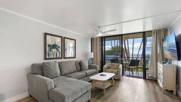 A modern living room with a grey sectional sofa, a white coffee table, and a patterned armchair. A ceiling fan is visible, and the room has large windows with a view of the outdoors.
