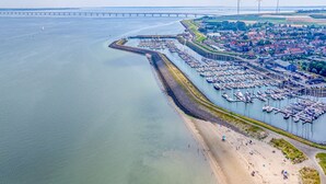 Luftaufnahme eines Küstenhafens mit zahlreichen dort vertäuten Booten, einem Sandstrand und einer Brücke im Hintergrund.