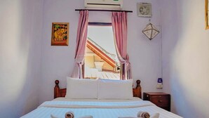 A hotel room with a bed, pillows, a nightstand, a wall-mounted air conditioner, a window with pink curtains, and a decorative wall hanging.
