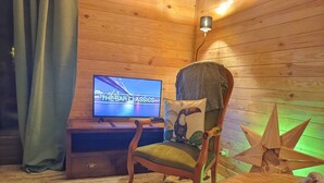 A wooden cabin interior with a television displaying 'THE BAR CLASSICS', a wooden chair with a cushion, and a small table with a plant.