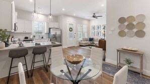A modern kitchen with white cabinets, a dining area with a glass table, and a living room with a sofa and decorative plates on the wall. - Modern Magnolia Cottage | Pool & Gym Access (Magnolia)