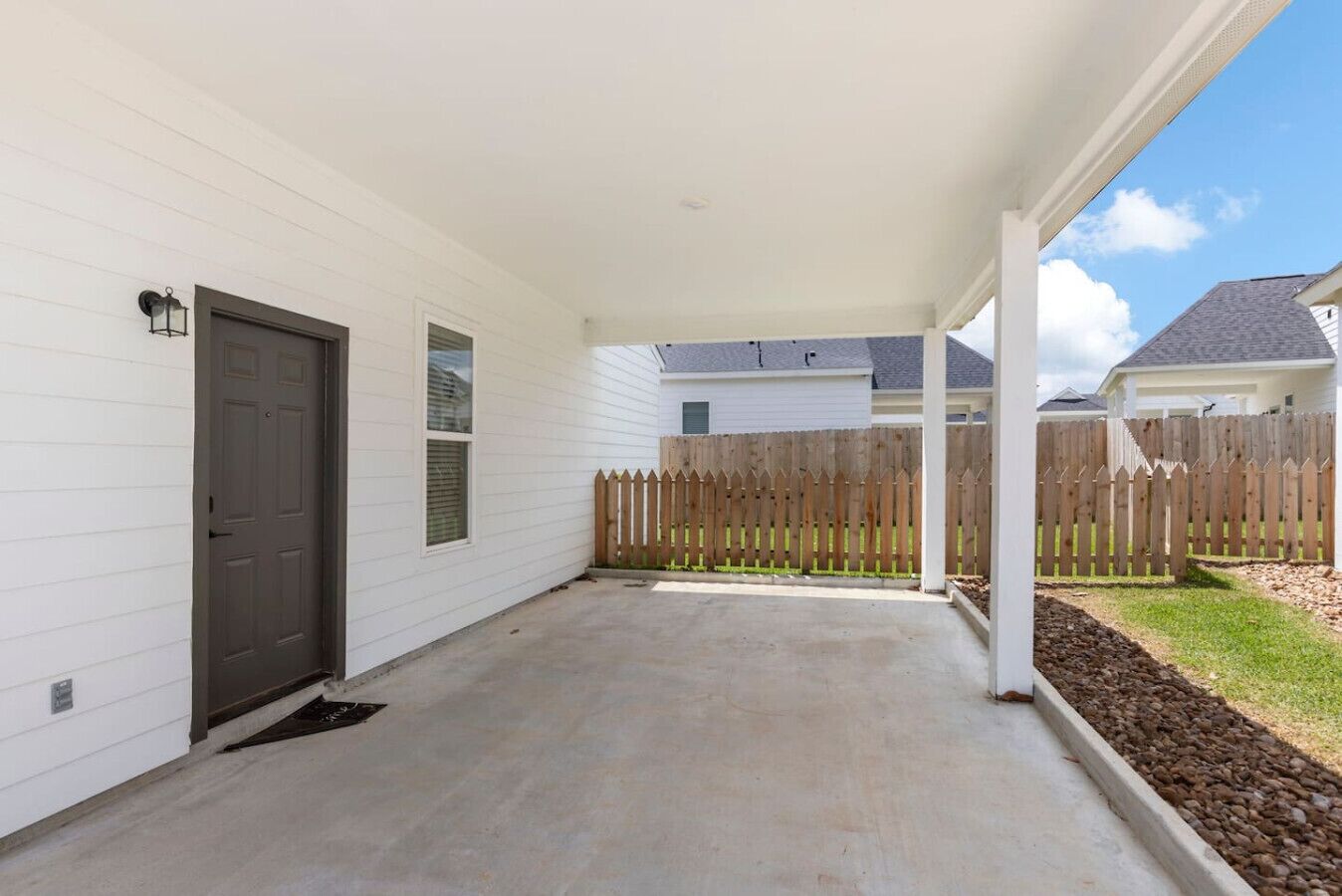 A covered patio with a concrete floor, a wooden fence, and a white house in the background.
