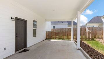 A covered patio with a concrete floor, a wooden fence, and a white house in the background.