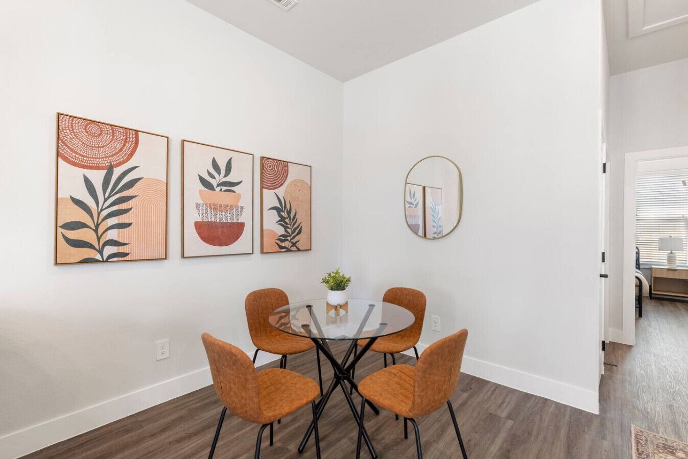 A dining area with a round glass table, four orange chairs, and three framed artworks on the wall.