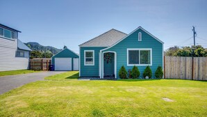 A two-story house with a front yard, a garage, and a wooden fence. - Walk to Gold Beach: Charming Cottage w/ Yard! (Gold Beach)
