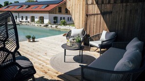 Une terrasse en bois avec des sièges d’extérieur, une piscine et un bâtiment avec un toit rouge en arrière-plan.