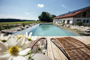 Une terrasse en bois avec deux fauteuils de jardin en osier, donnant sur une piscine rectangulaire, le tout entouré d’une clôture en bois et d’un bâtiment dont le toit est rouge.