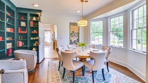 A dining room with a wooden table set for six, surrounded by chairs, a large window, and a bookshelf filled with books and decorative items. - ‘Wremy House’ 5 Blocks to Main St in Starkville! (Starkville)