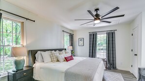 Un dormitorio con cama, mesitas de noche, ventilador de techo y ventanas amplias con cortinas.