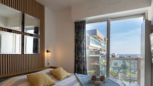 Ein modernes Schlafzimmer mit einem großen Bett, einem Spiegel, einem Nachttisch mit Lampe und einem Balkon mit Blick auf Gebäude und einen klaren Himmel.