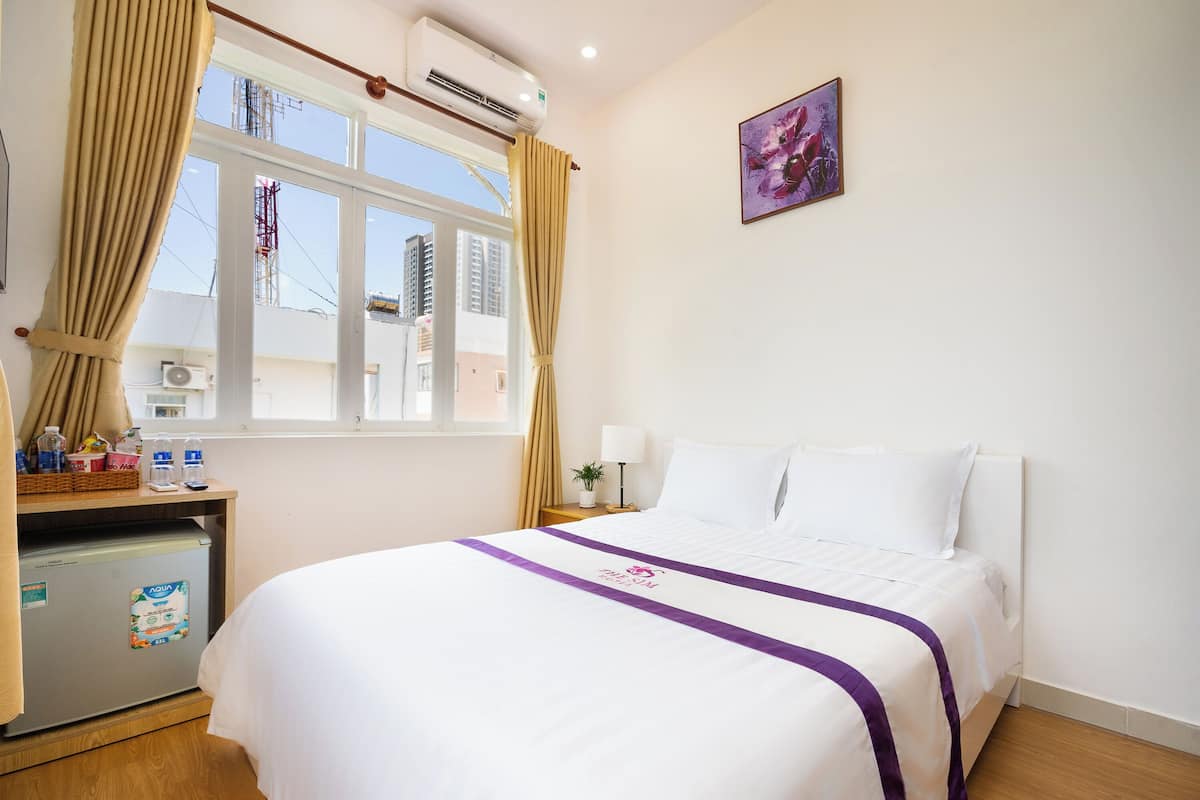 A hotel room with a bed, a desk with a refrigerator, a window with curtains, and a wall-mounted picture.
