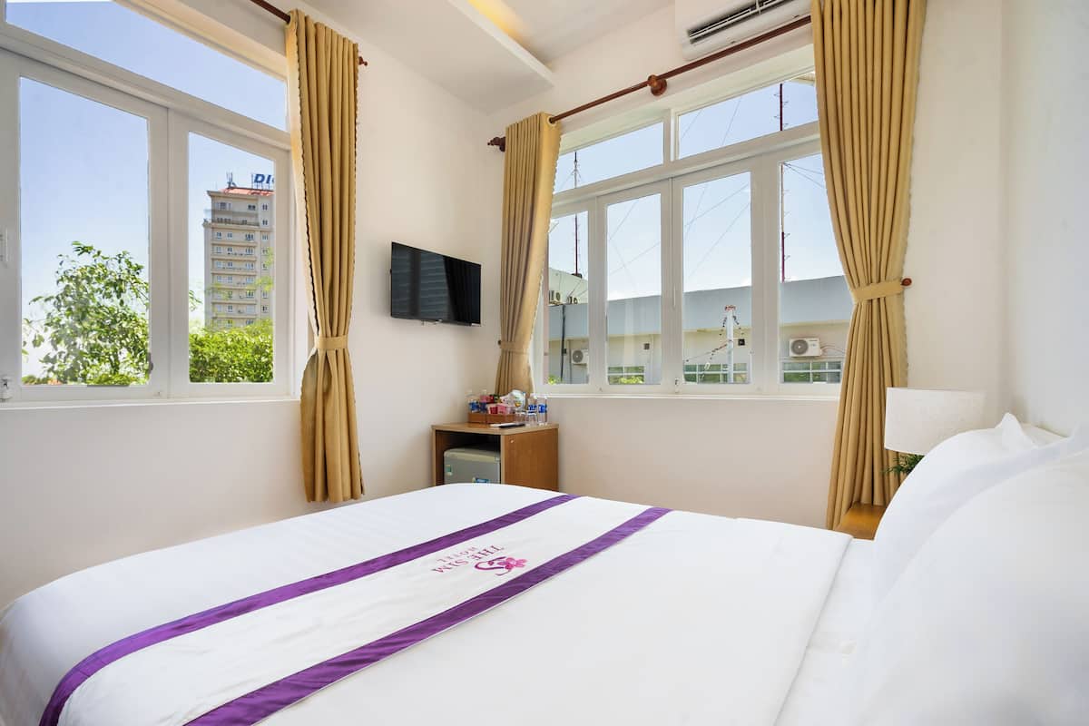 A hotel room with a bed, a TV mounted on the wall, a desk with a lamp, and a window with curtains.