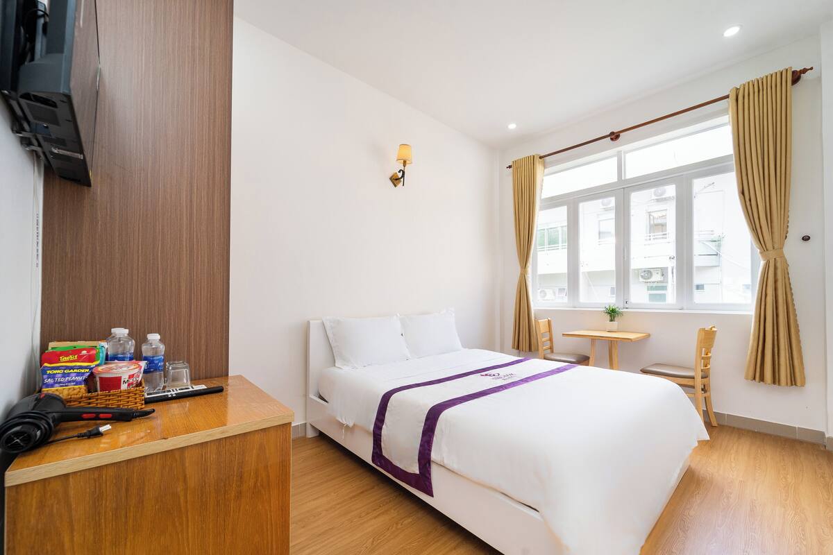 A hotel room with a bed, a desk with toiletries, a small table, and a window with curtains.