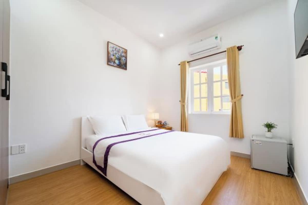 A hotel room with a single bed, a bedside table with a lamp, a small air conditioning unit, a window with curtains, and a wall-mounted air conditioning unit.