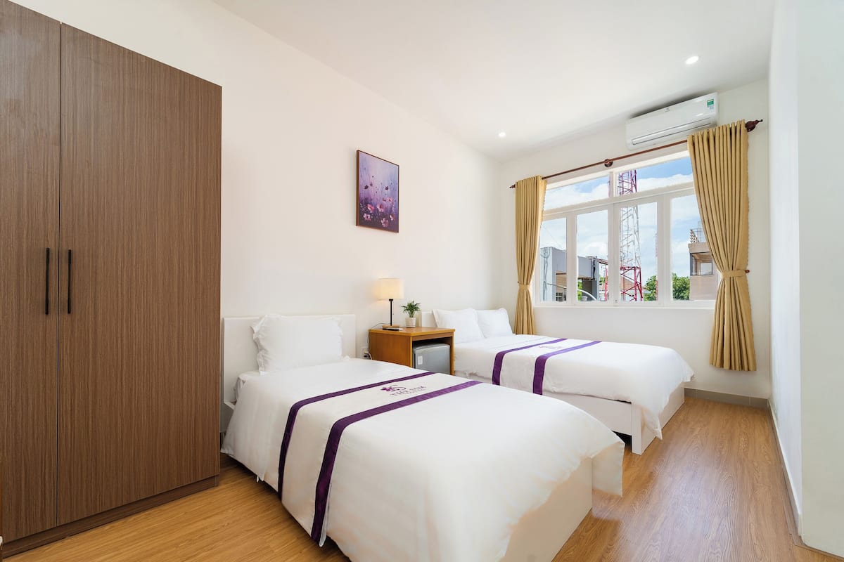A hotel room with two beds, a wardrobe, a bedside table with a lamp, a window with curtains, and an air conditioning unit.