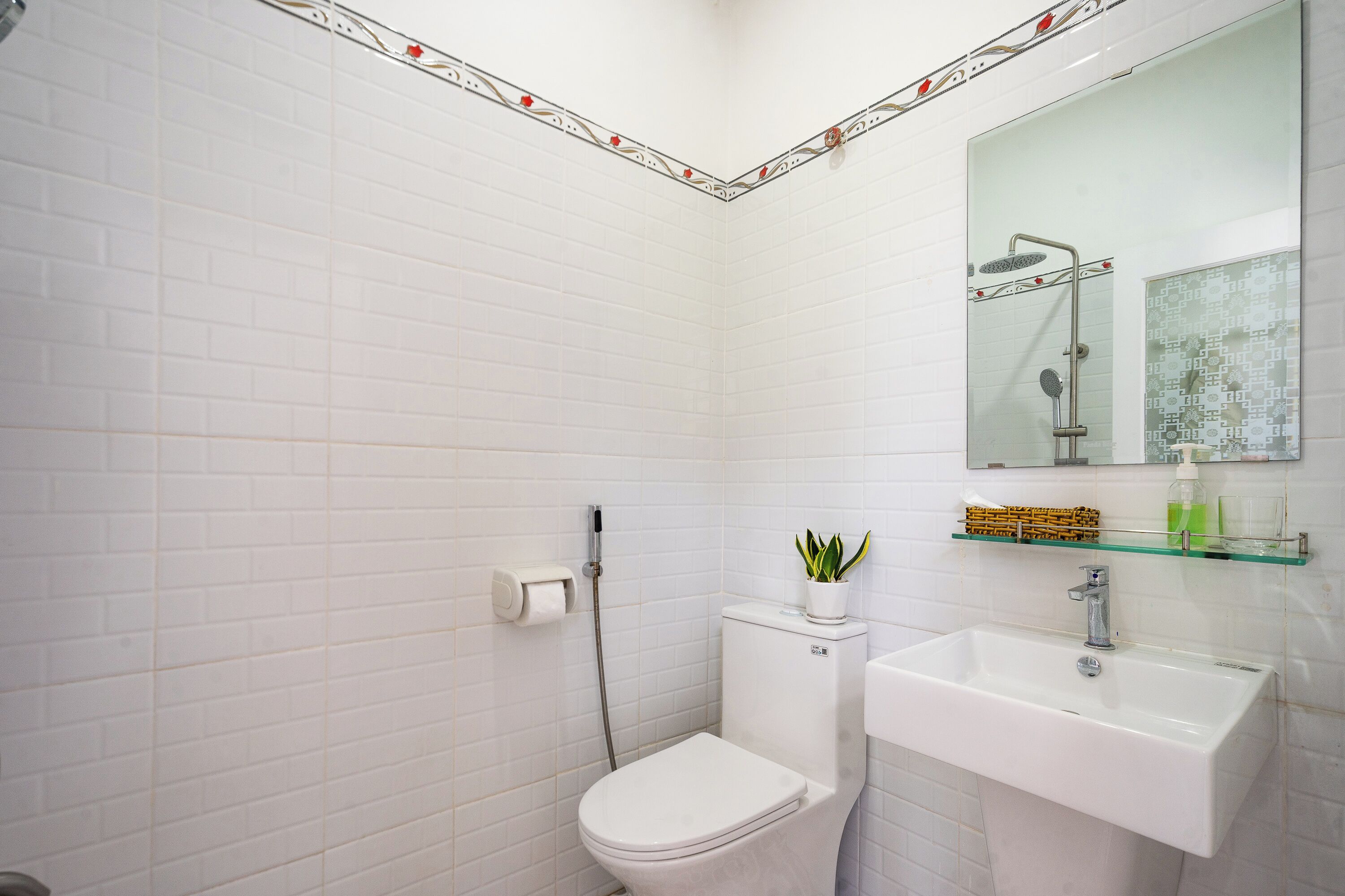 A bathroom with white tiled walls, a white toilet, a glass shelf with toiletries, and a white sink.